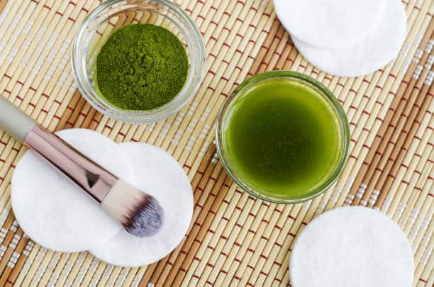 A jar of green face mask, a bowl of matcha powder, a makeup brush, and cotton pads are arranged on a woven mat.