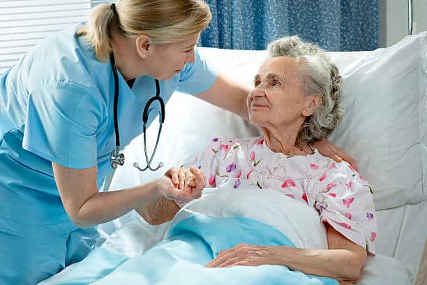 A nurse in blue scrubs holds the hand of an elderly patient who is lying in a hospital bed, offering comfort and support.