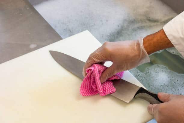 A person wearing gloves wipes a kitchen knife with a pink cloth over a white cutting board next to a sink filled with soapy water.
