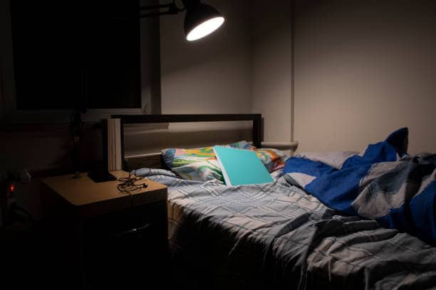 A dimly lit bedroom containing a bed with a colorful pillow, a blue throw blanket, and an open blue folder on it. A desk lamp illuminates the scene.