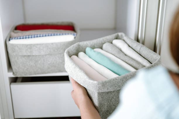 Person holding a gray fabric storage box filled with neatly folded clothes in front of a shelf with another similar box.