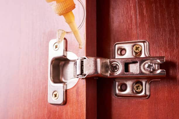A close-up of a metal hinge on a wooden door being lubricated with a clear liquid from a bottle with a narrow nozzle.
