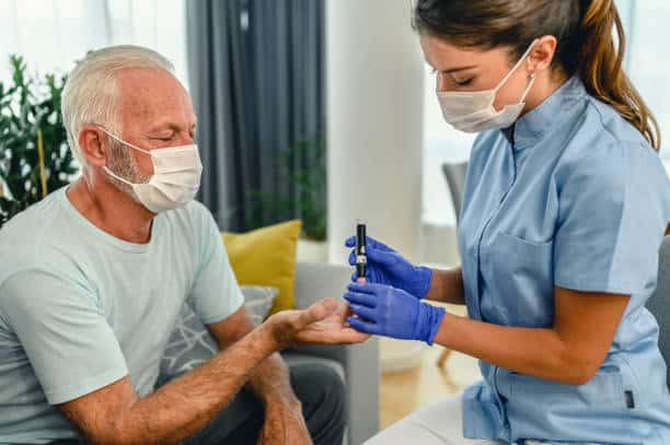 A healthcare worker in gloves and a mask checks blood glucose levels of an elderly man wearing a mask using a glucometer.