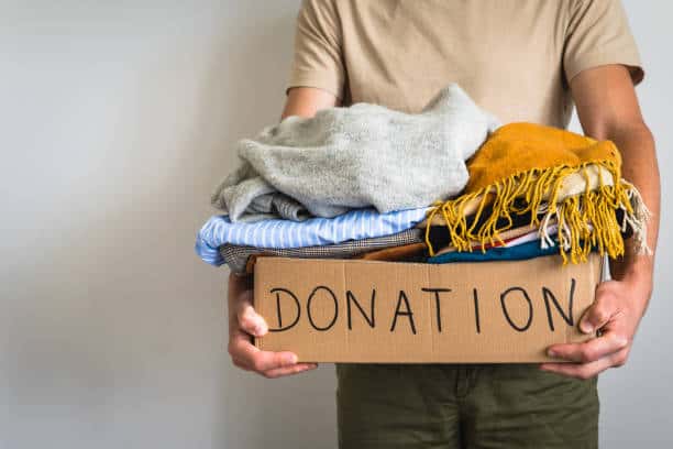 Person holding a cardboard box labeled "Donation" filled with folded clothes, including sweaters and a scarf.
