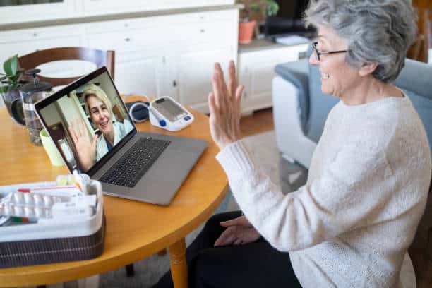 An elderly woman waves to a person on a video call on her laptop. A blood pressure monitor and medical supplies are nearby on the table.