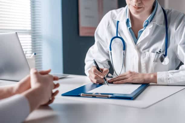 A doctor with a stethoscope writes on a clipboard while a patient sits across the desk.