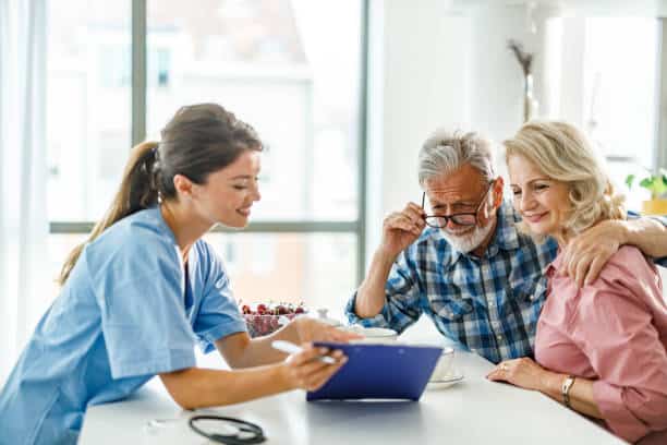 A nurse shows an elderly couple something on a clipboard at a table. The elderly man is adjusting his glasses while the elderly woman has her arm around him, both appearing engaged in the discussion.