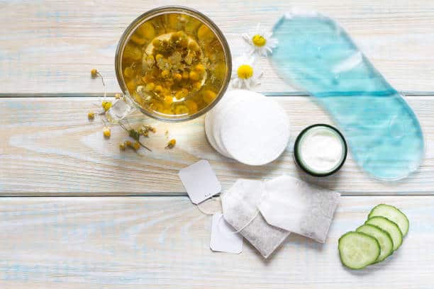 Overhead view of skincare items including a cup of chamomile tea, cotton pads, a gel eye mask, a jar of cream, tea bags, and cucumber slices on a light wooden surface.