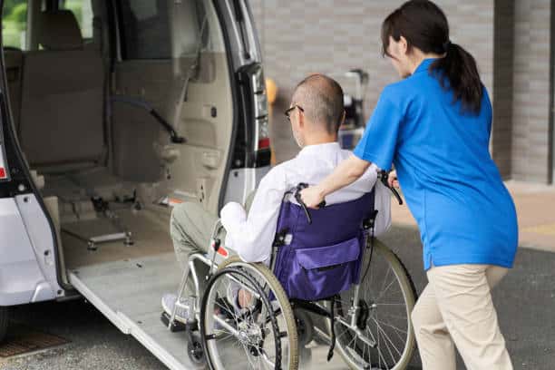 A caregiver assists a person in a wheelchair onto the ramp of a vehicle. The vehicle's back door is open and a ramp extends to the ground.
