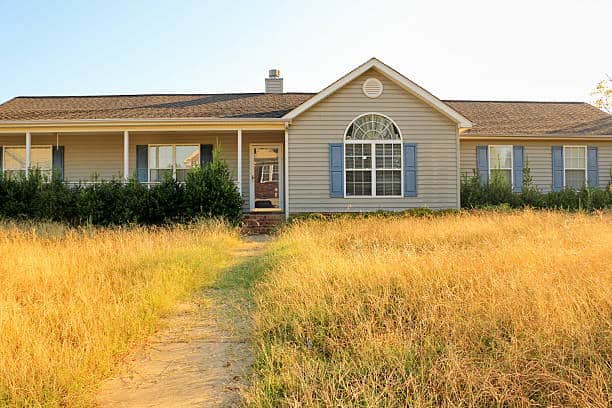 Single-story house with a front porch, blue shutters, and a large arched window. A path leads through tall, dry grass to the entrance.
