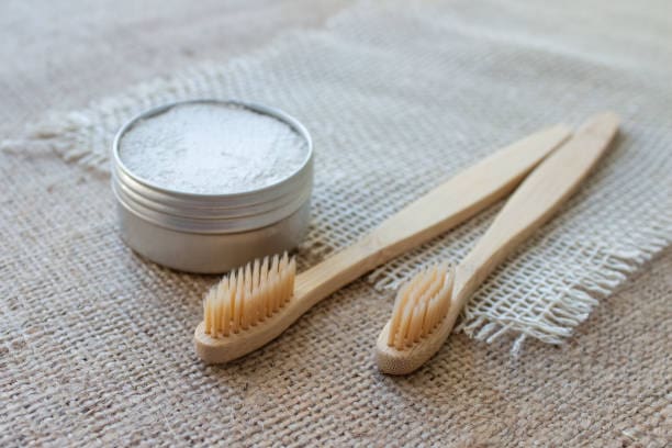 Two wooden toothbrushes and a round tin containing white powder lie on a burlap surface.