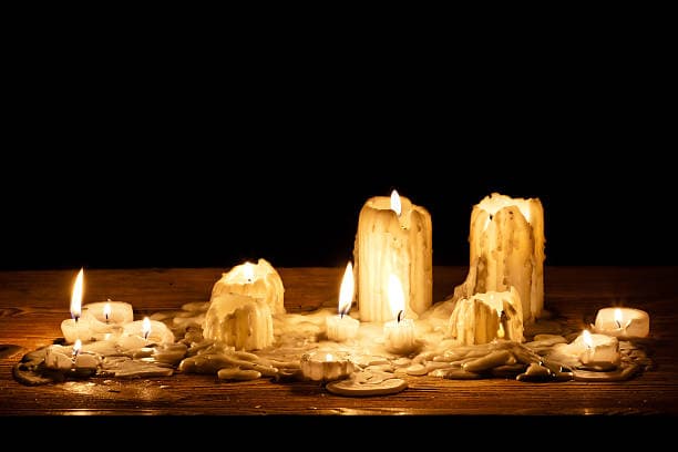 Lit candles of various sizes with melted wax pooling on a wooden surface against a dark background.