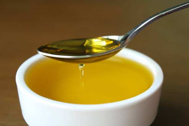 A spoon holding olive oil is shown above a white bowl filled with olive oil placed on a wooden surface.