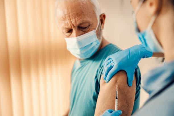 A man wearing a mask receives a vaccine shot in his arm from a healthcare worker wearing gloves and a mask.