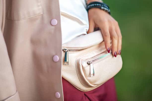 Close-up of a person wearing a beige fanny pack paired with a tan jacket. The person has painted nails and bracelets on their wrist.