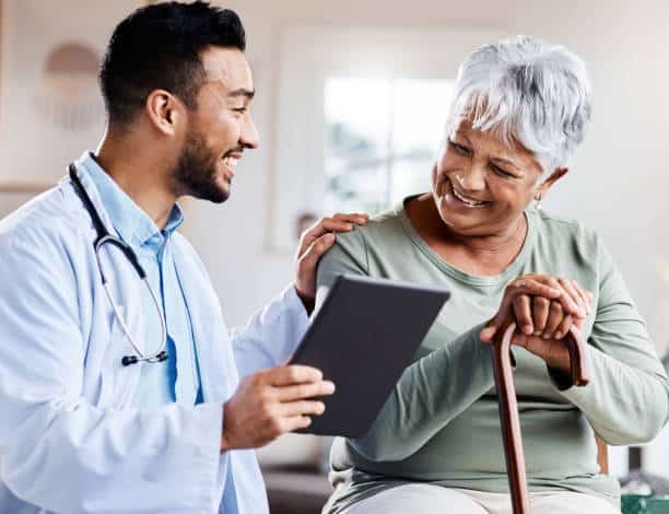 A doctor holds a tablet and talks with a seated older woman who is holding a cane. Both are smiling and appear engaged in their conversation.