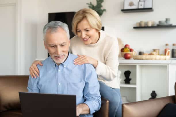 An elderly couple is smiling while using a laptop in a modern kitchen. The man is seated and working on the laptop, and the woman is standing behind him with her hands on his shoulders.
