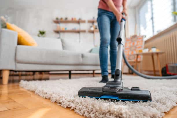 Person vacuuming a shag rug in a living room with a sofa and shelves in the background.