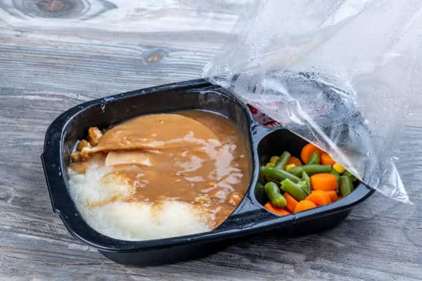 A microwave dinner with mashed potatoes and gravy, a meat patty, and mixed vegetables in a black plastic container with a partially removed plastic covering.