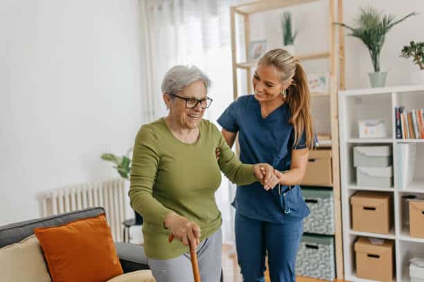 Elderly woman with a cane is assisted by a caregiver in a blue uniform inside a living room.