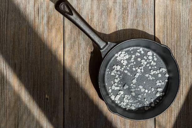 A cast iron skillet with granules of salt sits on a wooden surface with light and shadow patterns.