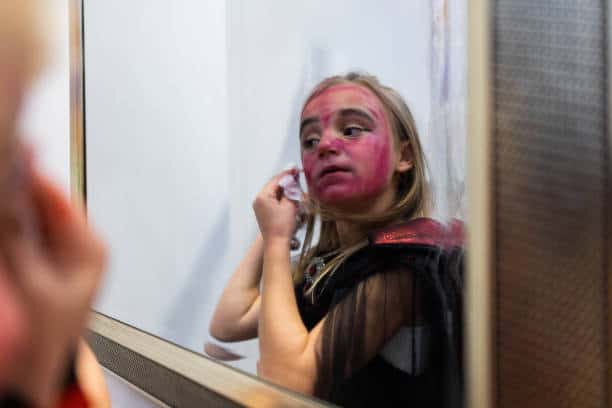 A person dressed in a costume looks into a mirror while wiping off red makeup from their face.