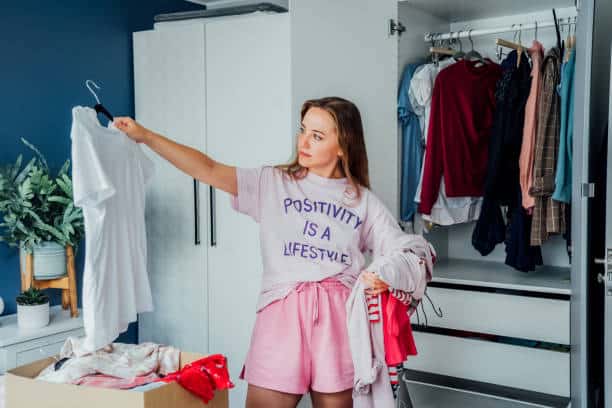 A woman stands in front of an open wardrobe, holding a white t-shirt on a hanger and wearing a light pink outfit. She appears to be organizing or considering her clothes.