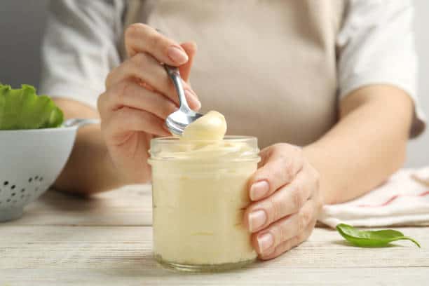 Person scooping mayonnaise from a jar with a spoon, with a bowl of lettuce in the background on a wooden table.