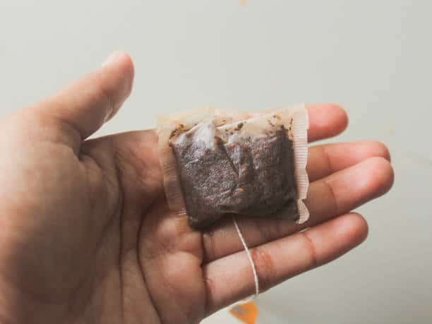 A hand holding a used tea bag with dark tea leaves visible inside the bag.