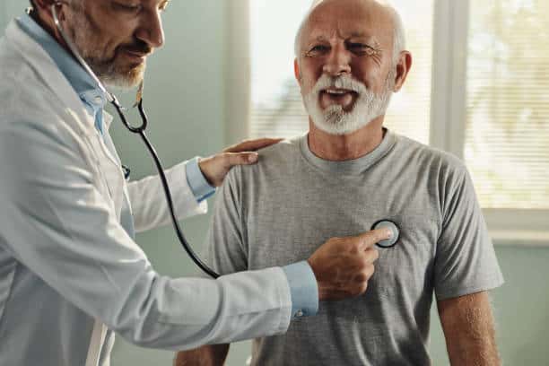 A doctor uses a stethoscope to examine an elderly male patient wearing a gray t-shirt, who is smiling.