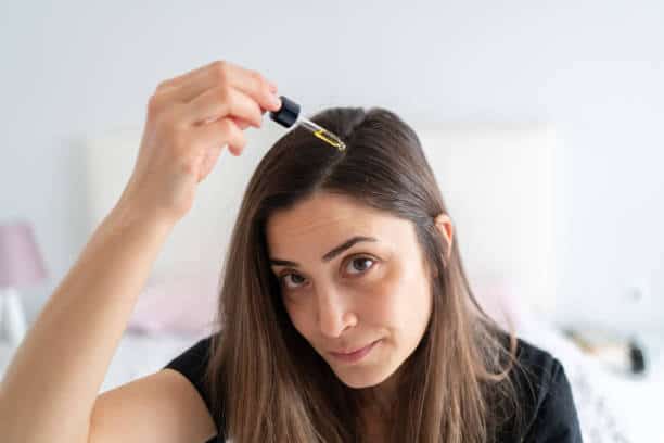 A woman with long brown hair applies oil to her scalp using a dropper.