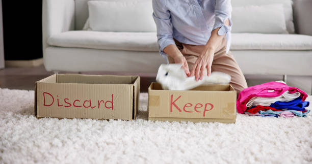 Person sorting clothes into two cardboard boxes labeled "Discard" and "Keep" in front of a couch.