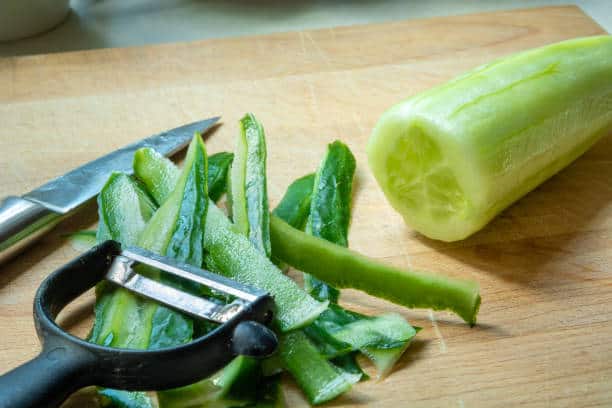 A peeled cucumber, vegetable peeler, and knife are placed on a wooden cutting board with cucumber peels.