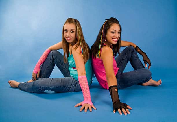 Two women are sitting back to back on a blue background. They are wearing jeans, colorful tops, and mesh fingerless gloves, and have small braids in their hair.