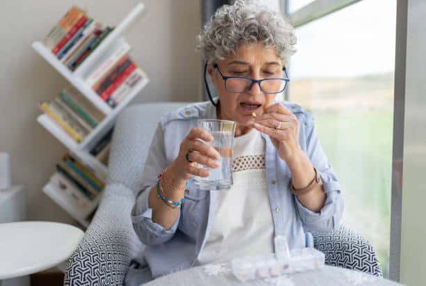 Elderly woman with glasses taking medication while holding a glass of water, seated beside a window with a bookshelf in the background.