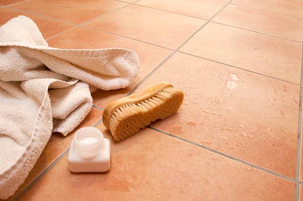 A floor cleaning scene with a scrub brush, a small bottle, and a white towel placed on a tiled floor that shows water droplets.