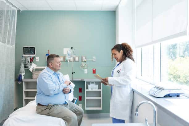 A man sits on a hospital bed while a doctor stands beside him, talking and holding a clipboard. The room is equipped with medical instruments and has a large window.