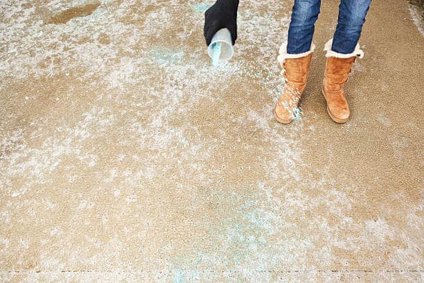 Person scattering ice melt on a snow-dusted surface, wearing jeans and tan snow boots adorned with fur.