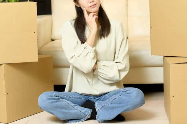 A person sits on the floor with legs crossed, surrounded by cardboard boxes, thinking or pondering.