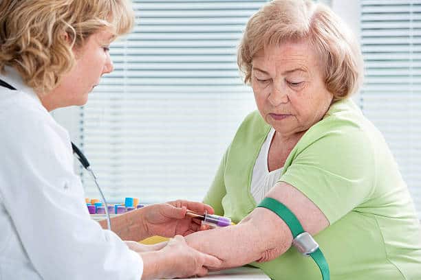 A healthcare professional draws blood from an elderly woman seated with a tourniquet on her upper arm.