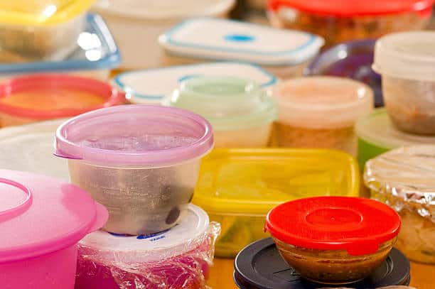 A variety of plastic food containers with different colored lids and some wrapped in plastic wrap are stacked together.