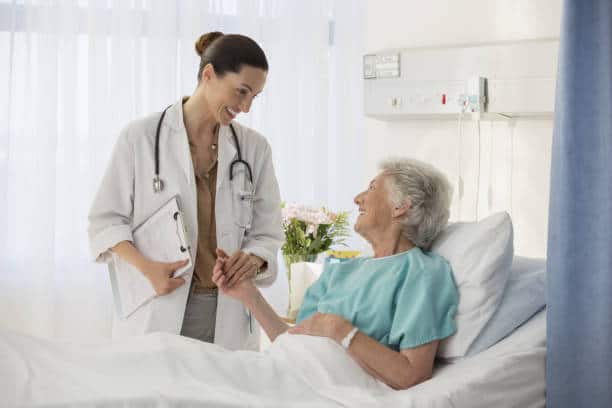 A doctor stands beside a hospital bed, holding a patient's hand while talking. The patient, an elderly woman, is lying in bed and smiling. Medical equipment is in the background.