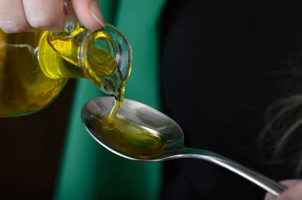 A hand pours olive oil from a glass bottle into a stainless steel spoon.