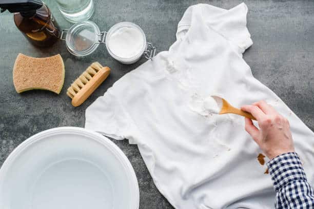 A person applies a cleaning substance to a white t-shirt stained with brown marks. The scene includes a brush, sponge, bowl of water, and cleaning supplies.