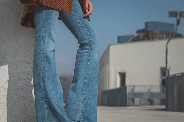 Person wearing flared blue jeans and a brown sweater stands outdoors near a concrete structure, with modern buildings in the background.