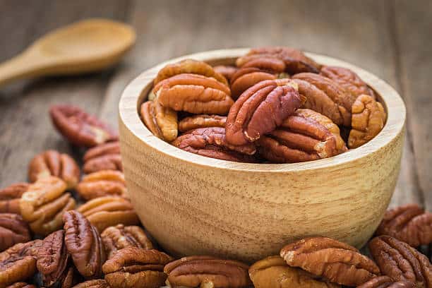 A wooden bowl filled with pecan nuts on a wooden surface, with some pecans scattered around the bowl.