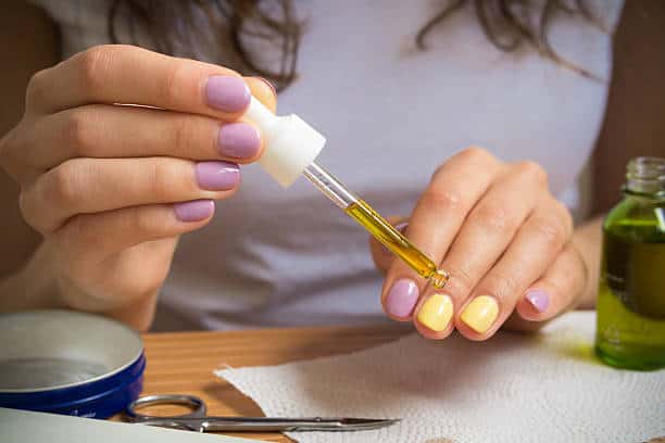 Close-up of hands using dropper to apply yellow oil onto fingers with pastel-painted nails. Scissors and a jar of oil are on the table.