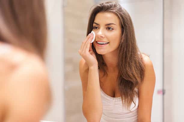 A woman looks in the mirror while using a cotton pad on her face in a bathroom.