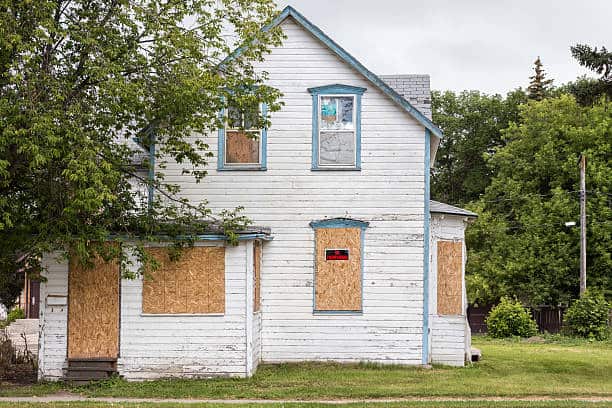 An old, two-story white house with boarded-up windows and a "For Sale" sign on the front window, surrounded by trees and grass.
