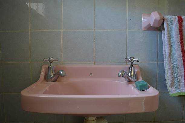 A pink ceramic sink with two silver faucets, soap on the right, and a red-striped towel on a pink holder against a tiled wall.